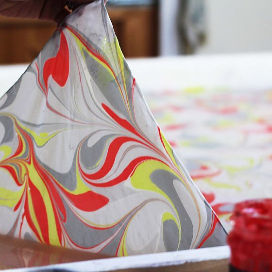 Silk being lifted off marbling tray to show pattern of red, grey, yellow and white swrils